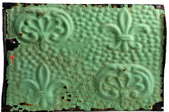 An old, green, tin, decorative tile with a fleur-de-lis design.