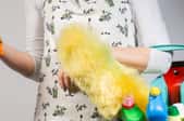 A woman holding a turquoise bucket full of cleaning supplies and a duster, and wearing an orange dusting mitt.