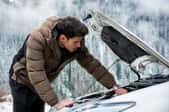 A man in a coat checking under the hood of a car in a winter setting.