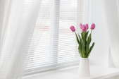 A window with white, sheer drapes and a vase of pink tulips sitting on the sill.