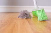 A green-bristled broom sweeping up a large dust bunny on a wood floor. 