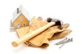 A small model of a house next to a hammer, pair of work gloves, and nails.