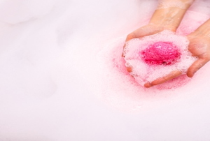 A pair of hands putting a pink bath bomb in a tub filled with water.