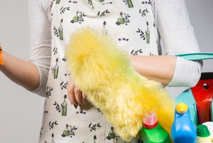 A woman holding a turquoise bucket full of cleaning supplies and a duster, and wearing an orange dusting mitt.