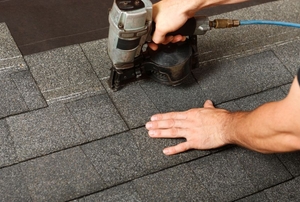 A man uses a nail gun to apply roof shingles.