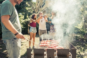 A BBQ with people and hot dogs cooking on the grill. 