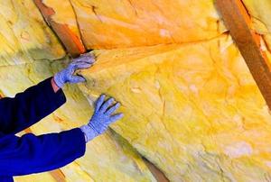 Blue-gloved hands installed a roll of yellow, foam insulation in an attic wall.