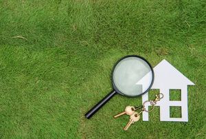 A paper house on grass with a magnifying glass and a set of house keys.