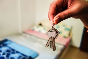 A set of house keys in the foreground with a bedroom in blurry focus in the background.