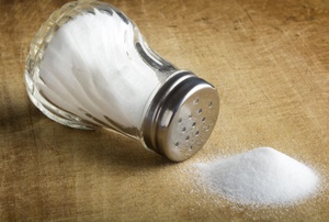 A pile of spilled salt on a wooden table from a shaker that is over turned.