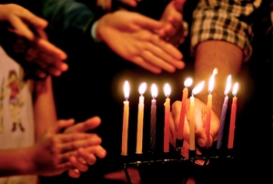 A menorah with lit candles and people's hands around it.