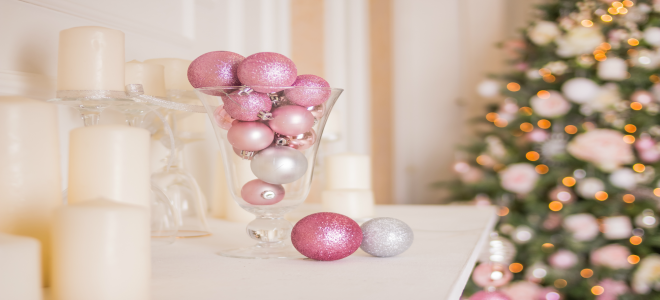 A vase of pink ornaments on a mantel with a Christmas tree in the background.