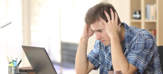 A man sitting in front of a laptop with his hands on his head worrying about something. 