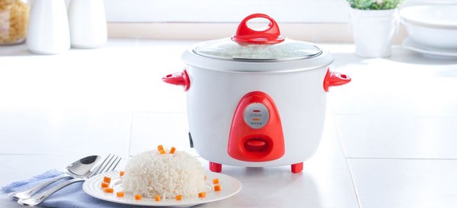 A small white and red rice cooker sitting on a counter beside a dish of prepared white rice.