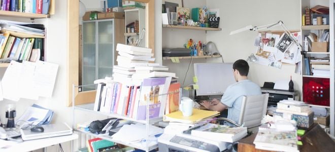 person working in cluttered home office with stacks of books