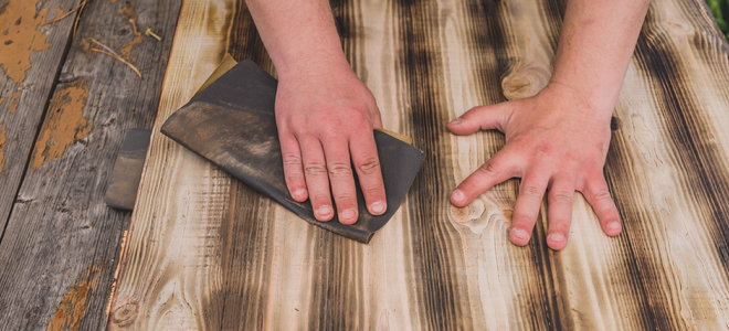 hands with sandpaper sanding down a dark wooden surface