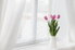 A window with white, sheer drapes and a vase of pink tulips sitting on the sill.