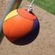 An orange tetherball hanging from a rope on a galvanized pole surrounded by dirt. 