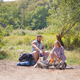 A couple sitting around a campfire during the day with green trees in the background.