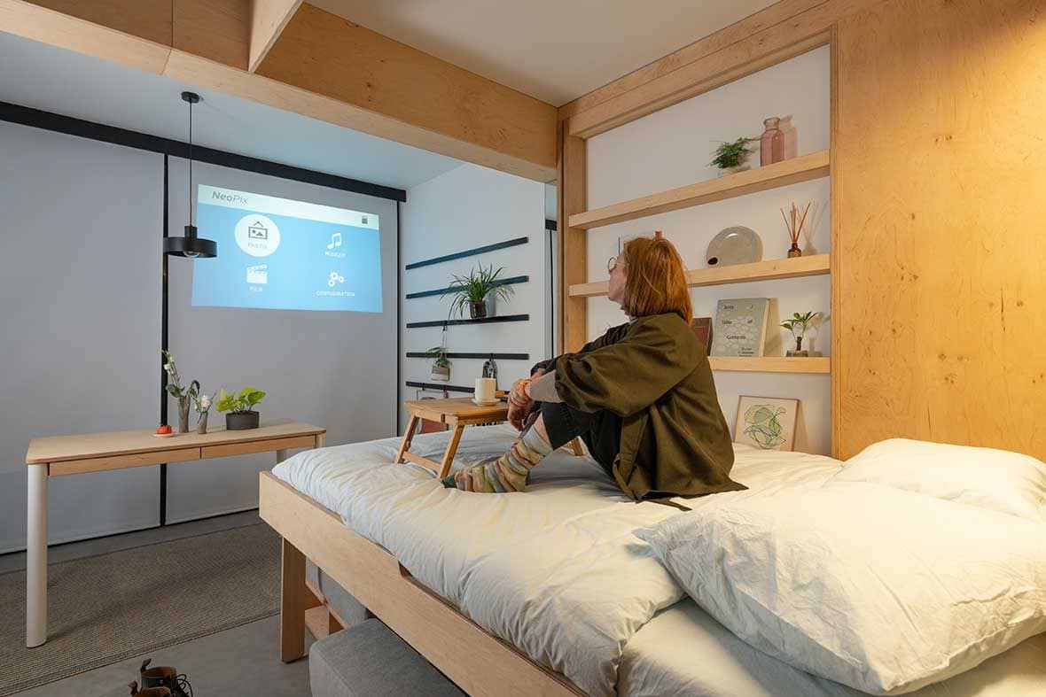 Woman watches TV in the cozy bedroom area of her Bouyges Immobilier stackable apartment module.
