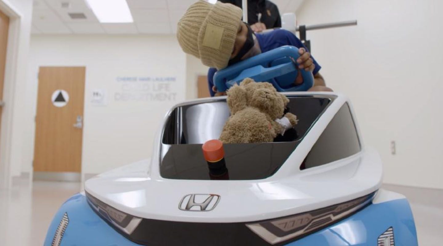 Sleepy child patient rides through the hospital in Honda's mini Shogo electric car with a teddy bear in the front.