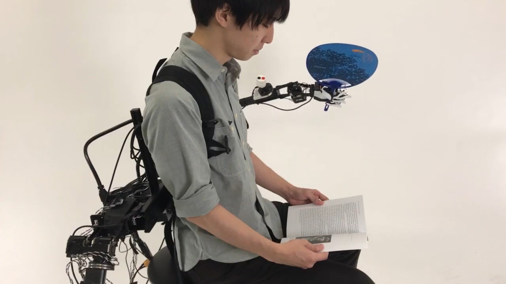 Man reads a book with his real arms while using the ISL robo-arms in Playback mode to swing a ping-pong paddle.
