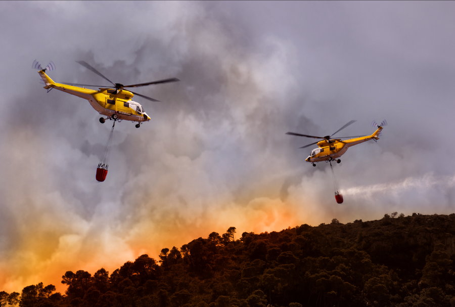 Two helicopters drop water buckets into the heart of blazing wildfires. 
