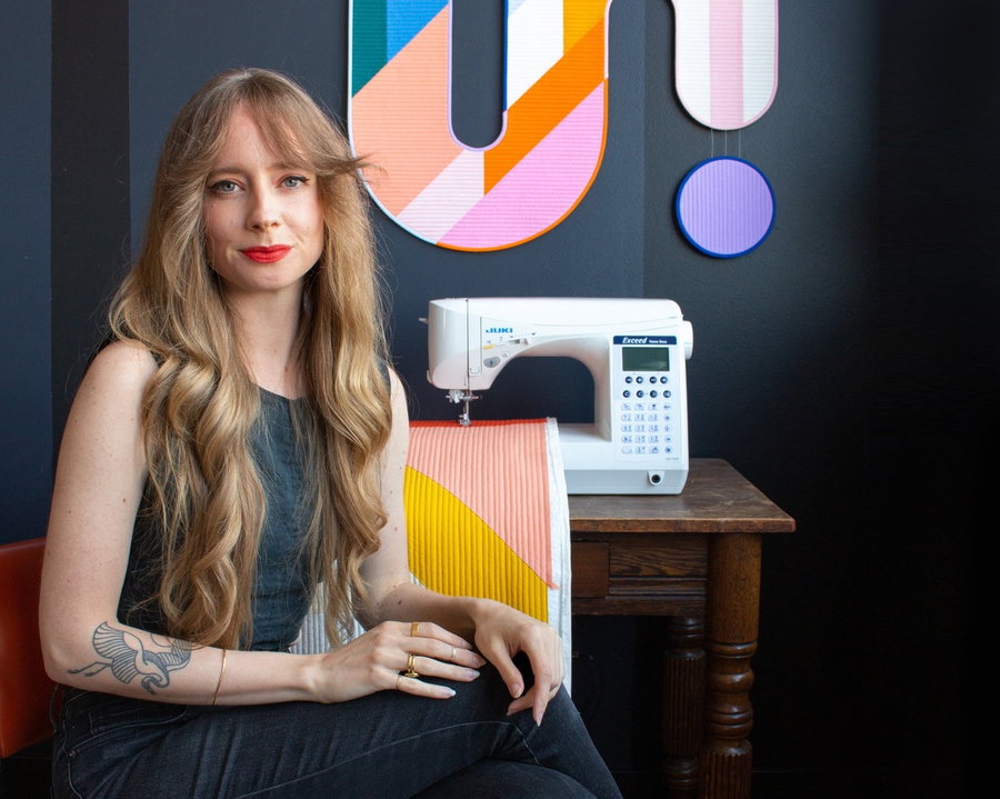 Emily Van Hoff sits in front of her sewing machine, above which one of hangs one of her quilted color block creations.