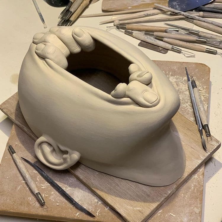 Surreal ceramic by Johnson Tsang shows hands reaching up from a stretched out face.
