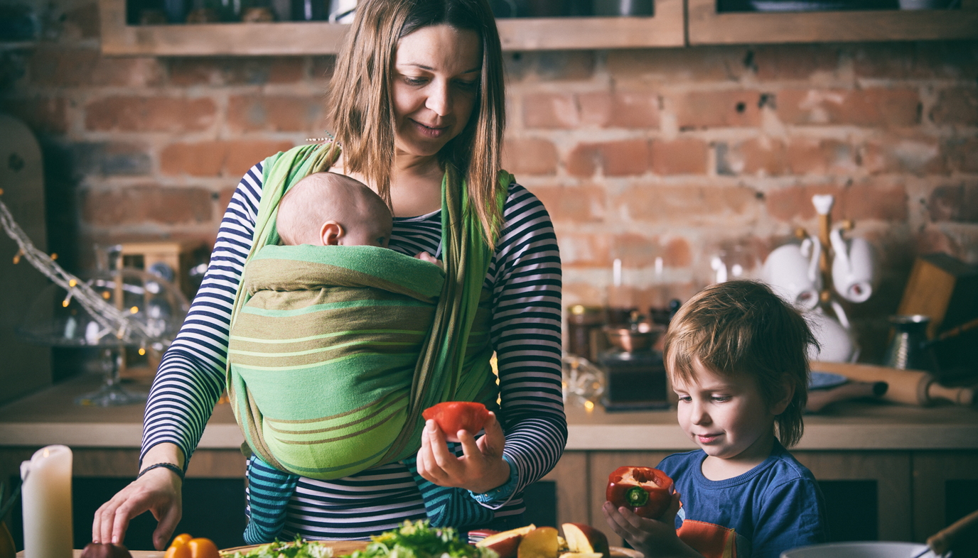 mom carrying baby in baby wrap while  preparing fruit with daughter