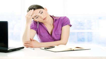 A woman sits at her desk with her eyes closed and a hand on her forehead. 