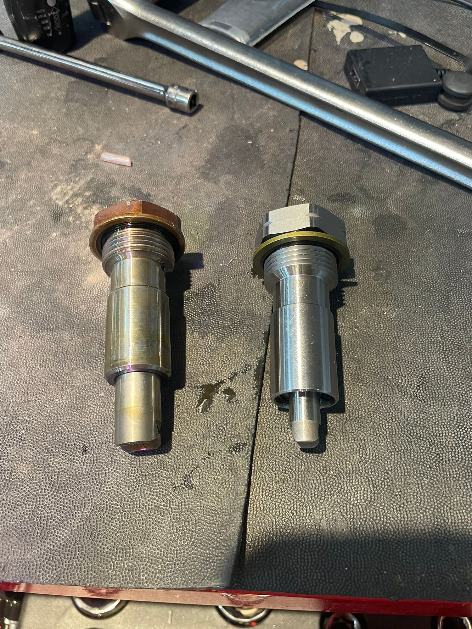 The defective OEM tensioner is on the left, the new is on the right. DO NOT activate before installing! It is normal for the shaft to slide in and out a few millimeters. The oil fill hole is near the washer.