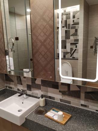 Large shower room with lit vanity mirrors and a set of vanity items (Chinese mouthwash, toothbrush/paste, q-tips, shower cap).